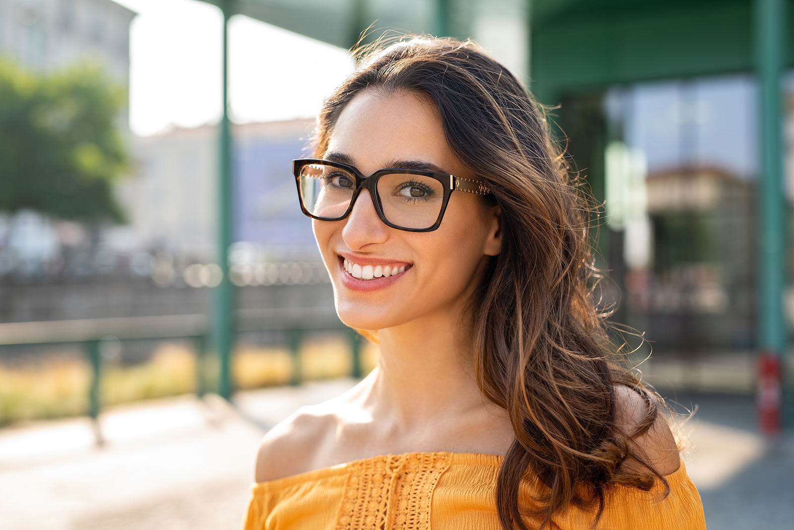 Portrait of carefree young woman smiling and looking at camera with urban background. Cheerful latin girl wearing eyeglasses in the city. Happy brunette woman with long hair and spectacles smiling.