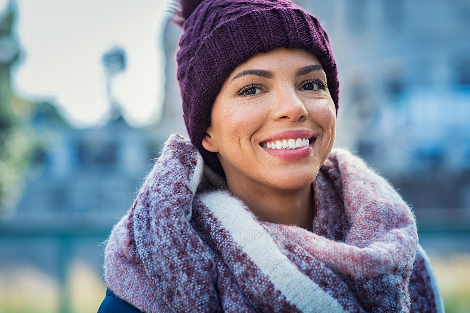 Closeup face of a young happy african woman enjoying winter wearing scarf and cap. Smiling brazilian girl looking at camera in a cold day. Multiethnic woman with knitted bordeaux hat and woolen scarf.
