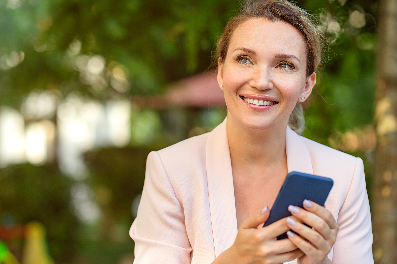 Close-up portrait of a smiling woman with smartphone on the street. Happy businesswoman is using phone, outdoors. Cheerful businesswoman in a jacket with cell phone in park looking away.